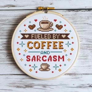 Op de afbeelding: Een borduurring met kruissteek met de tekst "FUELED BY COFFEE AND SARCASM". Het ontwerp bevat koffiekopjes, harten en decoratieve kruissteekelementen in bruin, rood en goud. De ring staat tegen een witte houten achtergrond.