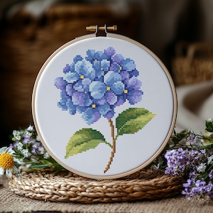Puede incluir: Un bastidor de bordado circular de madera que contiene tela blanca con un patrón de punto de cruz de una flor de hortensia azul y morada. La flor presenta pequeños centros amarillos, un tallo marrón y dos hojas verdes en estilo pixelado.
