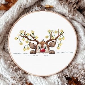 May include: A cross-stitch embroidery hoop featuring a reindeer design. The reindeer has brown antlers adorned with yellow lights and a red nose. The design is set against a white background, and the hoop is made of wood.