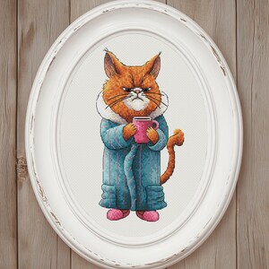 May include: Cross-stitch art of an orange cat with an angry expression, wearing a blue robe and pink slippers, holding a pink mug. The artwork is framed in a white oval frame, set against a wooden background.