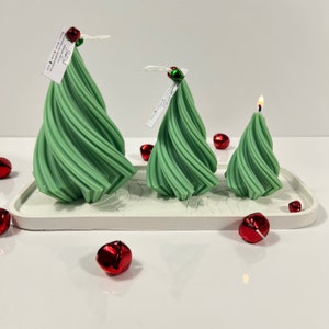 May include: Three green spiral Christmas tree candles of varying sizes on a white tray with red jingle bells scattered around.