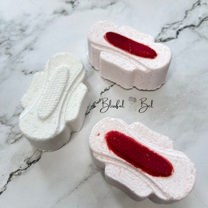May include: Three white bath bombs shaped like sanitary towels. Two of the bath bombs have a red centre, while the third is all white.