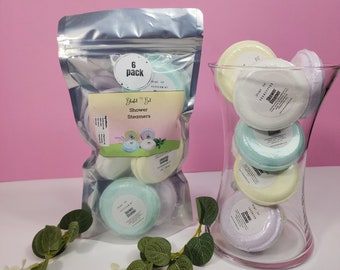 May include: A 6-pack of shower steamers in a silver resealable bag. The bag has a white circle with the text "6 pack" in black. The shower steamers are in a glass vase. The steamers are round and have different colours and scents. The scents are Lavender, Eucalyptus, Peppermint, and Tranquility.