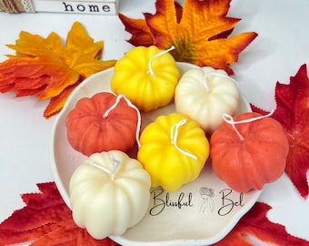 Puede incluir: Un conjunto de velas con forma de calabaza en tonos amarillo, naranja y crema, dispuestas sobre un plato blanco. Las velas están rodeadas de hojas decorativas otoñales. El texto "Blissful Bel" es visible.