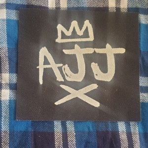 May include: A black fabric patch with white lettering that says "AJJ" with a crown above the letters and an "X" below.