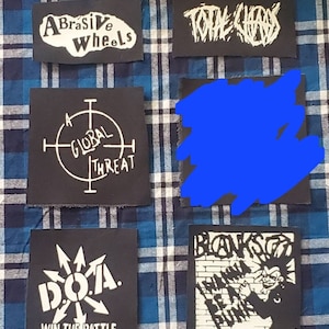 May include: A collection of eight black and white iron-on patches. The patches feature various designs, including a band logo, a target with the words "A Global Threat", a logo with the words "DOTA Win The Battle", and a logo with the words "Abrasive Wheels".