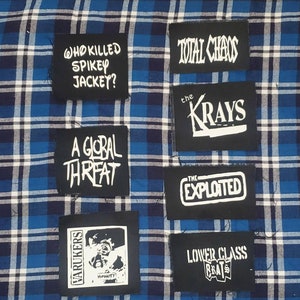 May include: A collection of eight black fabric patches with white text and graphics. The patches feature band names and slogans, including "Total Chaos", "The Krays", "The Exploited", "Lower Class Brats", "Varukers", "A Global Threat", "Who Killed Spiky Jacket?" and "Humanity".