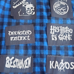 May include: A blue and black plaid flannel shirt with various band patches sewn on. The patches include "Anti-Sect", "Higher Hero Is Gone", "Deviated Instinct", "Disclose", "Beethoven", and "Kaos".
