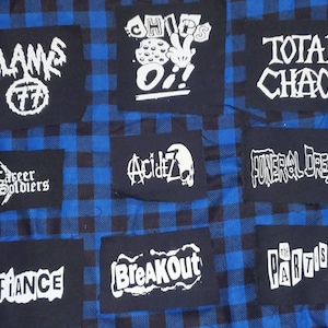 May include: A collection of black fabric patches with white text, featuring band names and phrases like "BLANKS 77", "TOTAL CHAOS", and "BREAKOUT". The patches are arranged on a blue and black plaid fabric background.