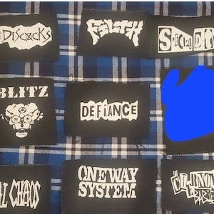 May include: A collection of black patches with white text and graphics. The patches feature band names like "BLITZ", "DEFiance", and "TOTAL CHAOS". The patches are displayed on a blue and white plaid background.