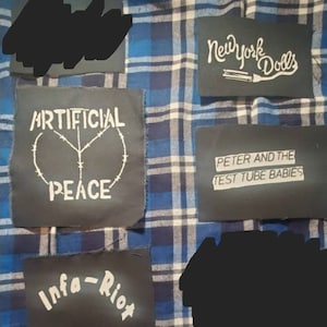 May include: A blue and white plaid shirt with several black patches sewn on. The patches have white text that reads "Artificial Peace", "New York Dolls", "Peter and the Test Tube Babies", and "Infra-Riot".