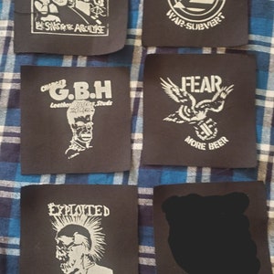 May include: A collection of eight black and white iron-on patches featuring punk rock band logos. The patches include logos for bands such as The Exploited, G.B.H., Fear, and The Mantas. The patches are all in a white and black color scheme and feature various band names and slogans.