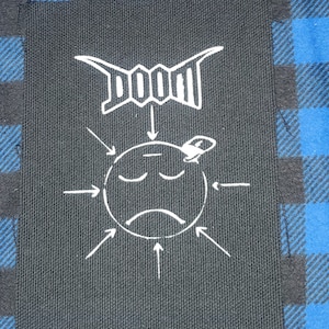 May include: A black fabric patch with white text and a cartoon face. The text says "DOOM" and an arrow points down to the cartoon face. The face is sad and has a small hat on top.