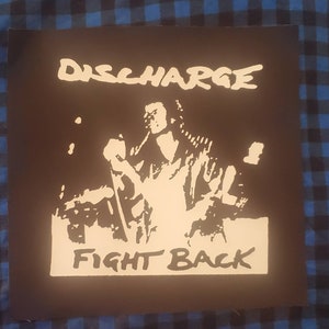 May include: A black and white image of a punk rock band called Discharge. The band is performing live on stage. The image has the band's name "Discharge" and the words "Fight Back" in white text.