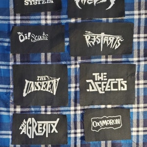 May include: A collection of eight black fabric patches with white text. The patches feature the names of various bands, including "One Way System", "Oi! Scouts", "The Restarts", "The Unseen", "The Defects", "The Agrestix", "Oxymoron", and "Twesy".