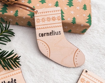 Könnte beinhalten: Weihnachtsstrumpf-Ornament aus Holz mit dem eingravierten Namen "Cornelius". Das Ornament hat ein dekoratives Muster oben und eine Lederschnur. Ein Geschenk mit grünen Weihnachtsbäumen ist im Hintergrund.
