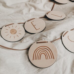Peut inclure: Une guirlande en bois avec un soleil, un arc-en-ciel et la lettre E. La guirlande est enfil&eacute;e sur un cordon brun et est parfaite pour une chambre d'enfant ou une cr&egrave;che.