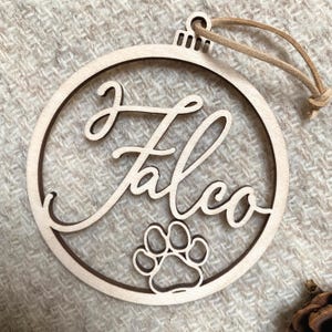 Könnte beinhalten: Ein hölzerner Weihnachtsbaumschmuck mit dem Namen "Falco" in Kursivschrift und einem Pfotenabdruck. Der Schmuck ist hellbraun und hat eine Schnur zum Aufhängen.
