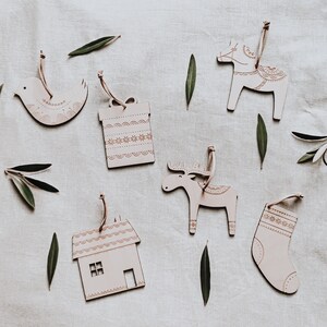 May include: Set of six wooden Christmas ornaments in various shapes, including a house, a reindeer, a stocking, a gift box, a bird, and a horse. The ornaments are painted white with intricate patterns in gold.