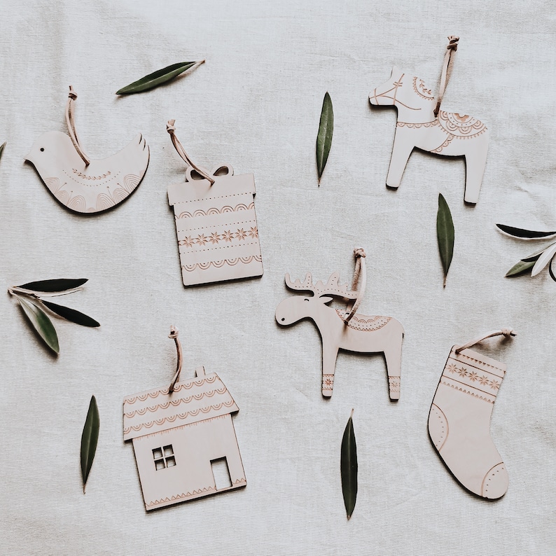 May include: Set of six wooden Christmas ornaments in various shapes, including a house, a reindeer, a stocking, a gift box, a bird, and a horse. The ornaments are painted white with intricate patterns in gold.