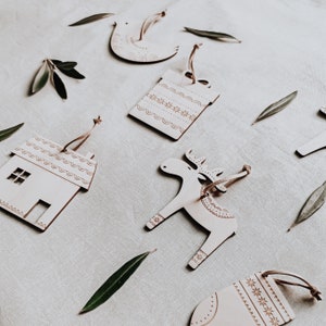 May include: A set of six wooden ornaments with a Scandinavian design. The ornaments are in the shape of a house, a reindeer, a bird, and three geometric shapes. The ornaments are painted white with a brown pattern.