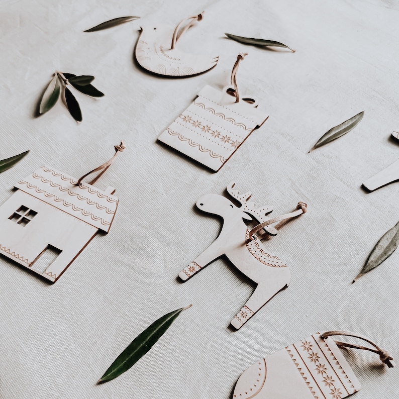 May include: A set of six wooden ornaments with a Scandinavian design. The ornaments are in the shape of a house, a reindeer, a bird, and three geometric shapes. The ornaments are painted white with a brown pattern.
