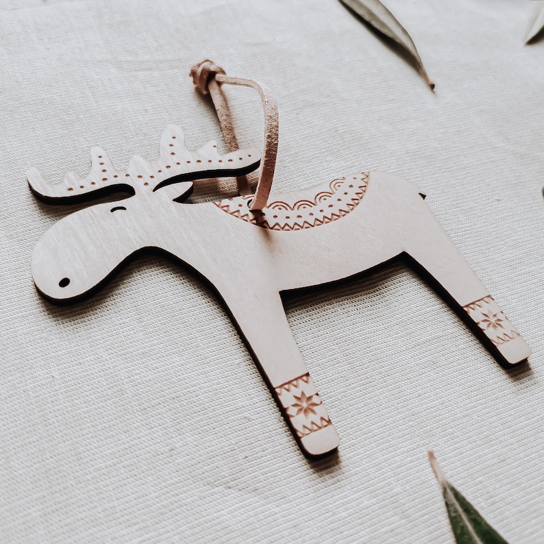 May include: A wooden reindeer ornament with a brown leather loop for hanging. The reindeer has a detailed design with a snowflake pattern on its legs and a decorative pattern on its back.