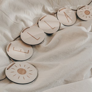 Peut inclure: Une banni&egrave;re en bois avec le nom "ELA" &eacute;crit en lettres blanches sur des pi&egrave;ces rondes en bois individuelles. La banni&egrave;re est enfil&eacute;e sur un cordon beige et comprend un motif de soleil &agrave; chaque extr&eacute;mit&eacute;.