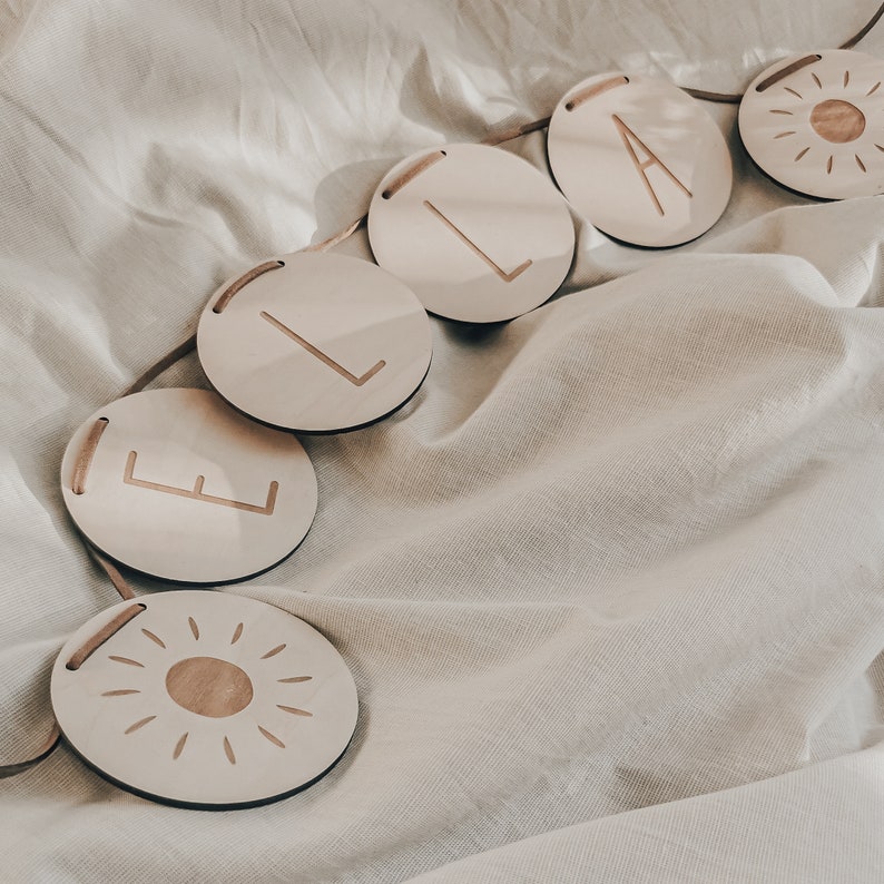 Peut inclure: Une banni&egrave;re en bois avec le nom "ELA" &eacute;crit en lettres blanches sur des pi&egrave;ces rondes en bois individuelles. La banni&egrave;re est enfil&eacute;e sur un cordon beige et comprend un motif de soleil &agrave; chaque extr&eacute;mit&eacute;.