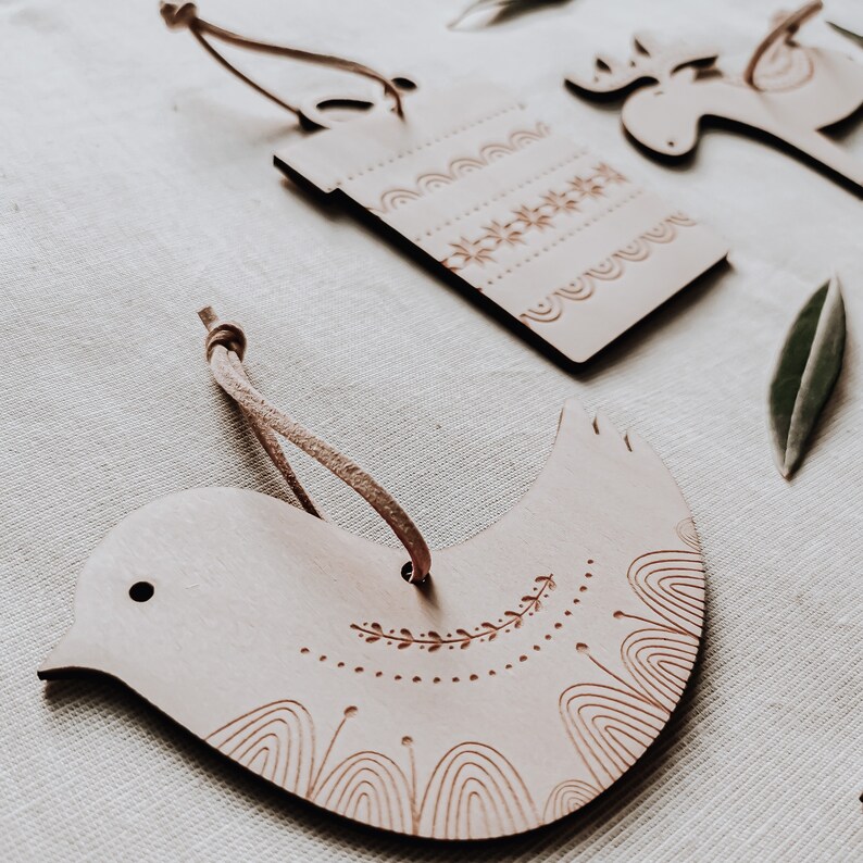 May include: Three wooden ornaments with a rustic, natural finish. The ornaments are shaped like a bird, a reindeer, and a gift box. Each ornament has a simple, hand-drawn design.