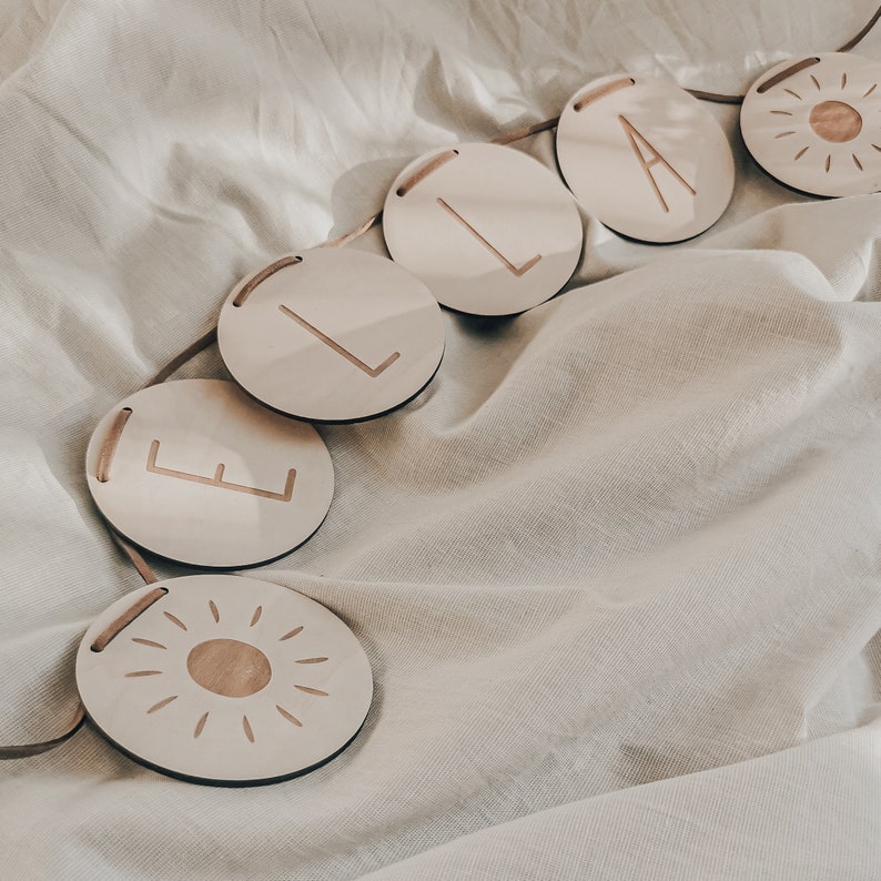 Peut inclure: Une banni&egrave;re en bois avec le mot "ELSA" &eacute;crit en lettres individuelles dans des cercles, avec un motif de soleil &agrave; chaque extr&eacute;mit&eacute; de la banni&egrave;re.