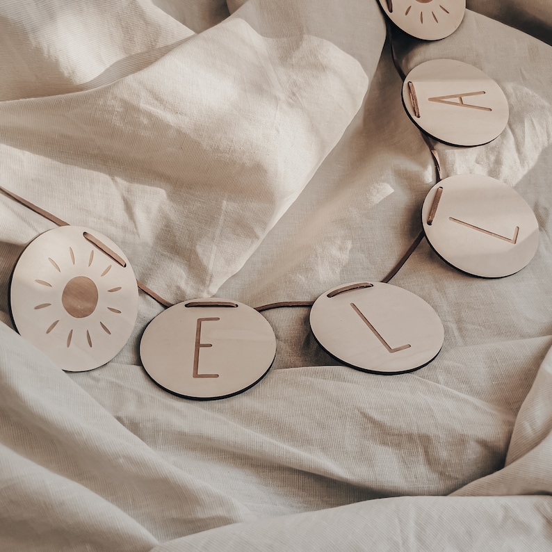 Peut inclure: Une guirlande en bois blanc avec un soleil et les lettres "LOVE" dans une police simple et moderne. La guirlande est enfil&eacute;e sur une ficelle et est suspendue sur un fond de tissu blanc.