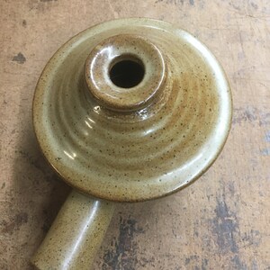 May include: A small, brown ceramic pot with a single handle and a round opening in the top. The pot has a speckled glaze and a slightly textured surface.