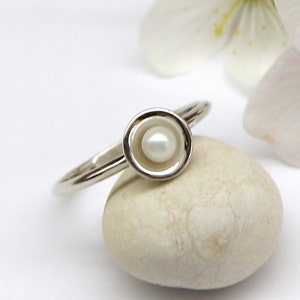 May include: A silver ring with a white pearl set in a circular bezel. The ring is resting on a white stone.
