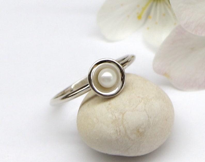 May include: A silver ring with a white pearl set in a circular bezel. The ring is resting on a white stone.