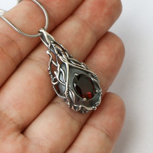 May include: A silver pendant necklace with a dark red gemstone set in a delicate, intricate silver design. The pendant is shaped like a teardrop and features a detailed, organic pattern.