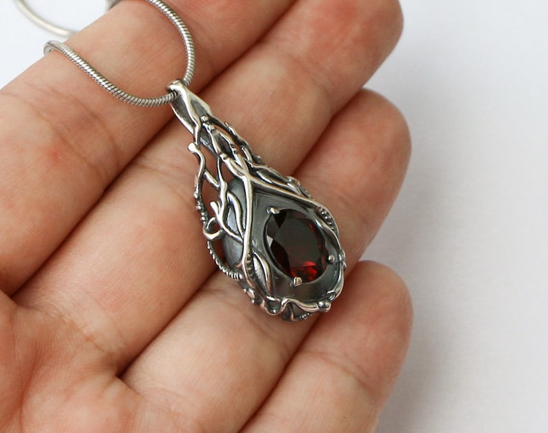 May include: A silver pendant necklace with a dark red gemstone set in a delicate, intricate silver design. The pendant is shaped like a teardrop and features a detailed, organic pattern.