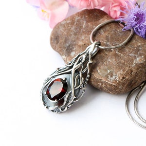 May include: A silver pendant necklace with a red gemstone set in a delicate, intricate design. The pendant is shaped like a teardrop and features a silver chain.