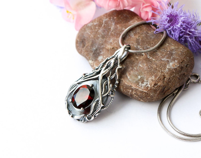 May include: A silver pendant necklace with a red gemstone set in a delicate, intricate design. The pendant is shaped like a teardrop and features a silver chain.