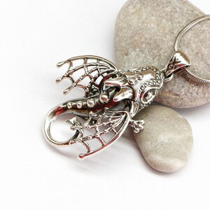 May include: Silver dragon pendant necklace with intricate details. The dragon is in a flying pose with its wings spread out. The pendant is attached to a silver chain.