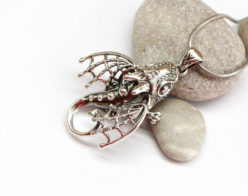 May include: Silver dragon pendant necklace with intricate details. The dragon is in a flying pose with its wings spread out. The pendant is attached to a silver chain.