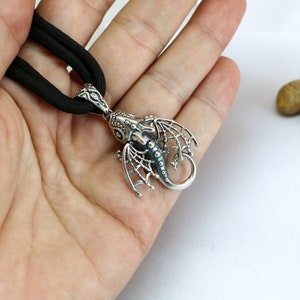 May include: A silver dragon pendant with intricate details, hanging from a black cord necklace.