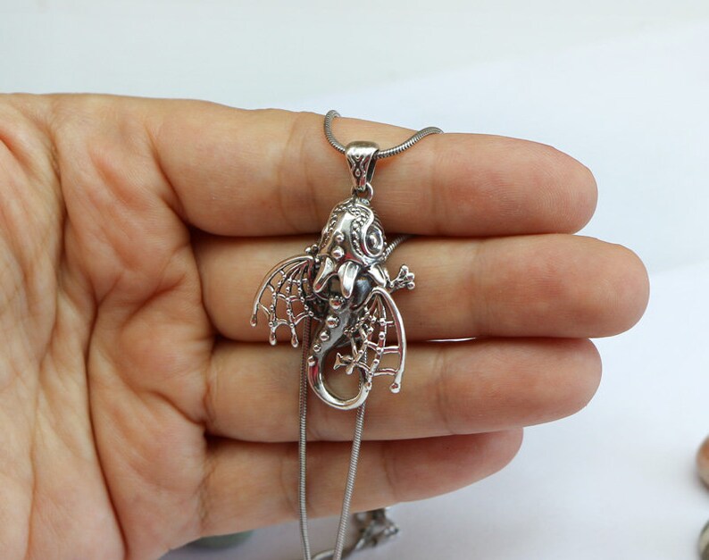 May include: Silver dragon pendant necklace with intricate details and a black stone in the center. The dragon is curled up with its wings spread out.