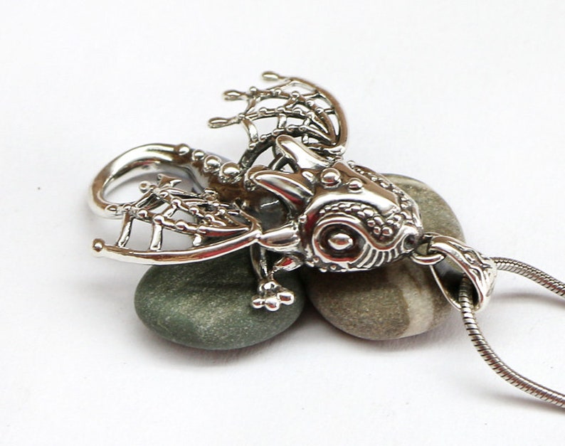 May include: A silver pendant featuring a detailed dragon design with intricate wings and a textured body. The pendant is hanging from a silver chain.