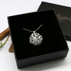 May include: A silver necklace with a round pendant featuring a delicate design of leaves. The necklace is displayed in a black box with the word "Queen" printed on the lid.