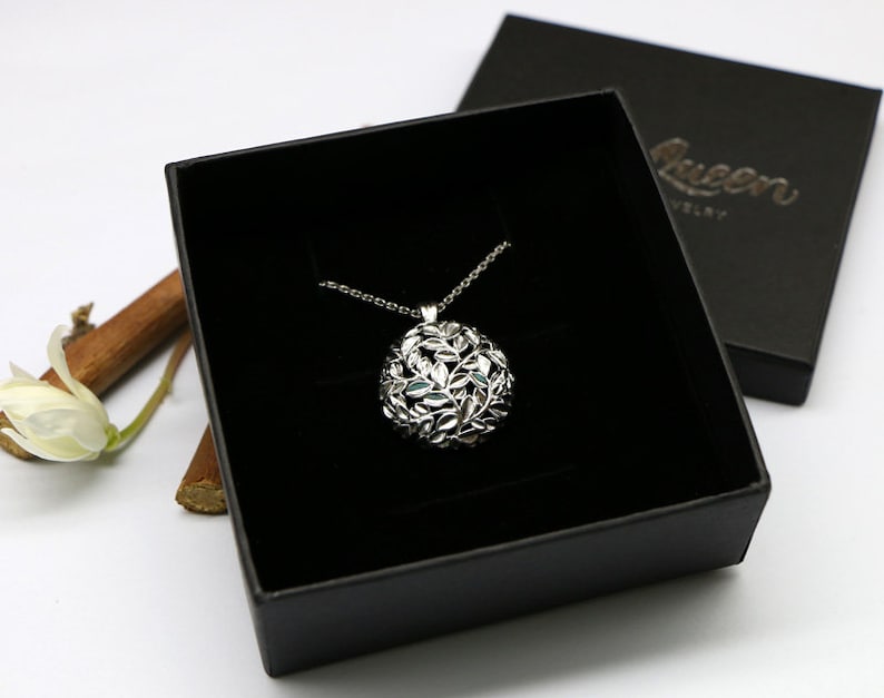 May include: A silver necklace with a round pendant featuring a delicate design of leaves. The necklace is displayed in a black box with the word "Queen" printed on the lid.