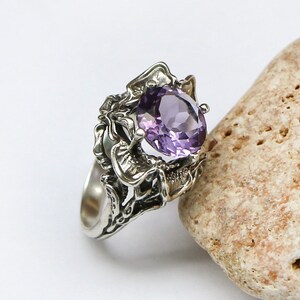 May include: A silver ring with an intricate design featuring a large, oval-shaped amethyst gemstone.