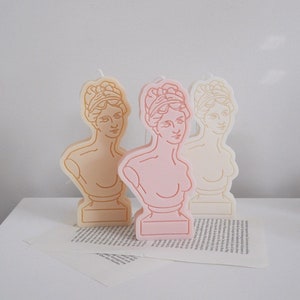 Peut inclure: Trois bougies décoratives en forme de buste de femme. Les bougies sont dans des tons beige, rose et blanc. Le buste est délimité par un dessin au trait en or, rose et blanc.