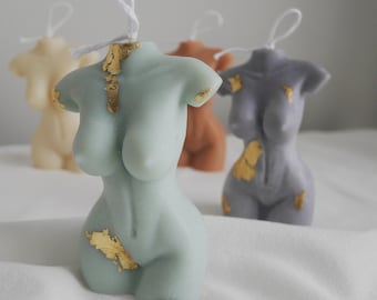 May include: A collection of torso-shaped candles in various colors, including a light green, tan, orange, and gray. Each candle is adorned with gold leaf accents and has a white wick. The candles are arranged on a white surface.