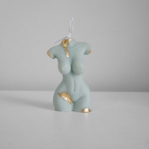 Peut inclure: Une bougie en forme de torse bleu clair avec des accents de feuilles d'or. La bougie a une m&egrave;che en ficelle et est pr&eacute;sent&eacute;e sur une surface blanche. Un objet de d&eacute;coration unique.